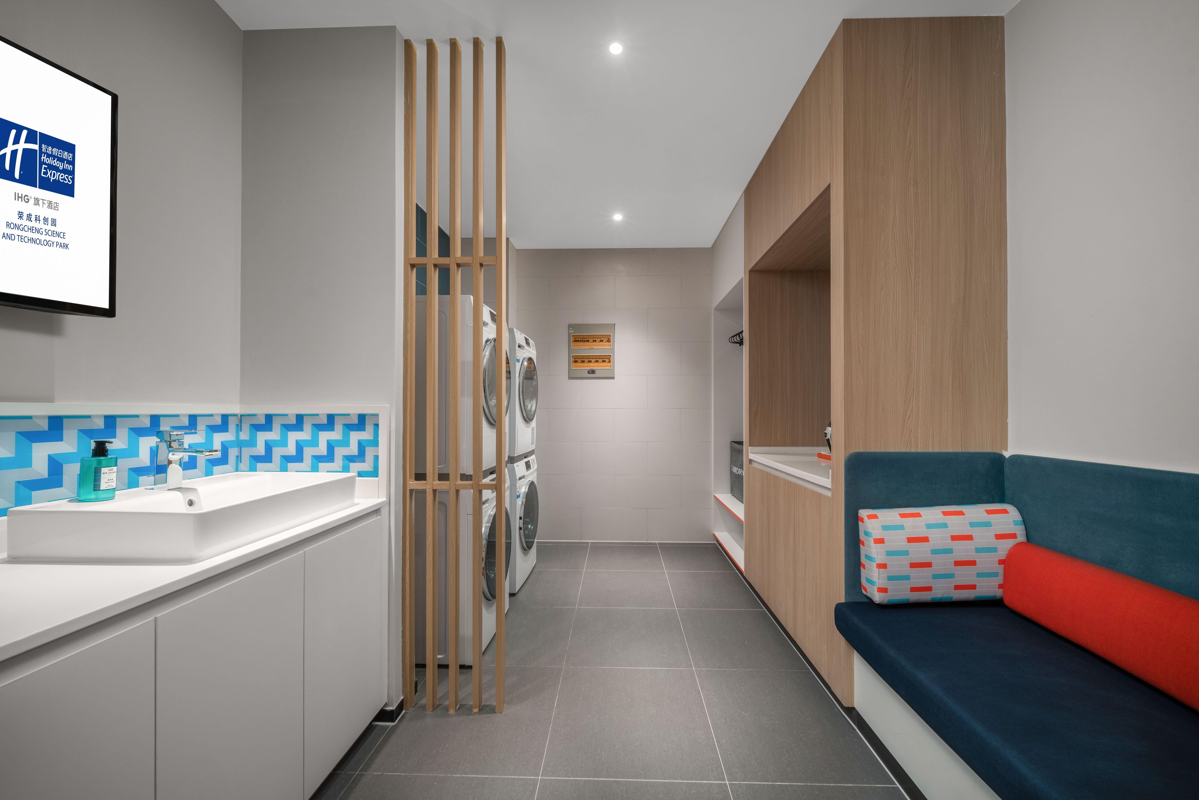 A modern hotel room with a bathroom, a washing machine, a sofa, and a wall-mounted TV.