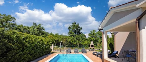 A rectangular swimming pool with a tiled deck, surrounded by lush greenery and a clear blue sky.