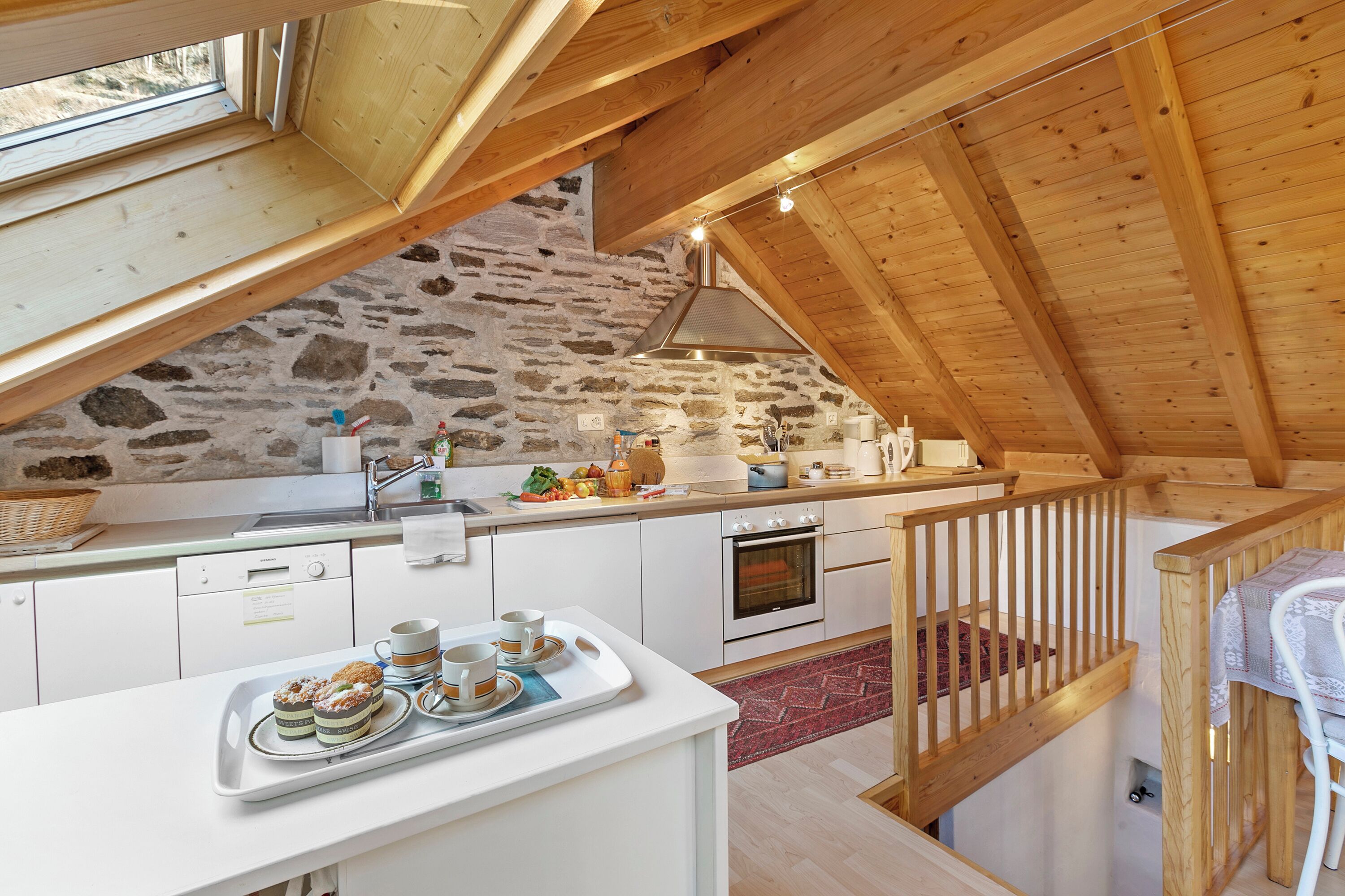 A rustic kitchen with stone walls, wooden beams, and a wooden staircase.