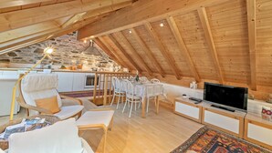 A cozy attic room with a wooden ceiling, a dining area with a table and chairs, a sofa, a TV, and a kitchen area with a stone wall.