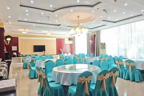 Une salle de banquet avec des tables rondes, des chaises de couleur turquoise ornées de nœuds beige, un lustre et une grande fenêtre.