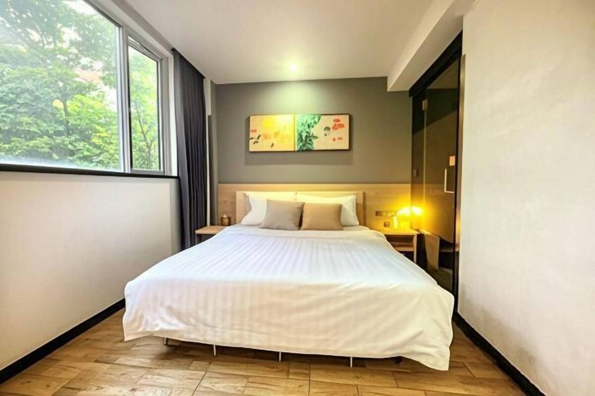 A bedroom with a large bed, a nightstand, a wall-mounted lamp, and a window with a view of trees.