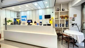 A reception desk with a computer, a monitor, and a plant. There is a bookshelf with books and decorative items. A table with chairs is visible in the foreground. - Hi Inn (Shanghai Jinshan Zhujing Jinlong New Street) (Shanghai)