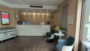 A hotel reception area with a marble counter, two blue chairs, a small table with a plant, and a trash bin. - Shankee Hotel (Qingdao Development Zone Xiangjiang Second Road) (Qingdao)