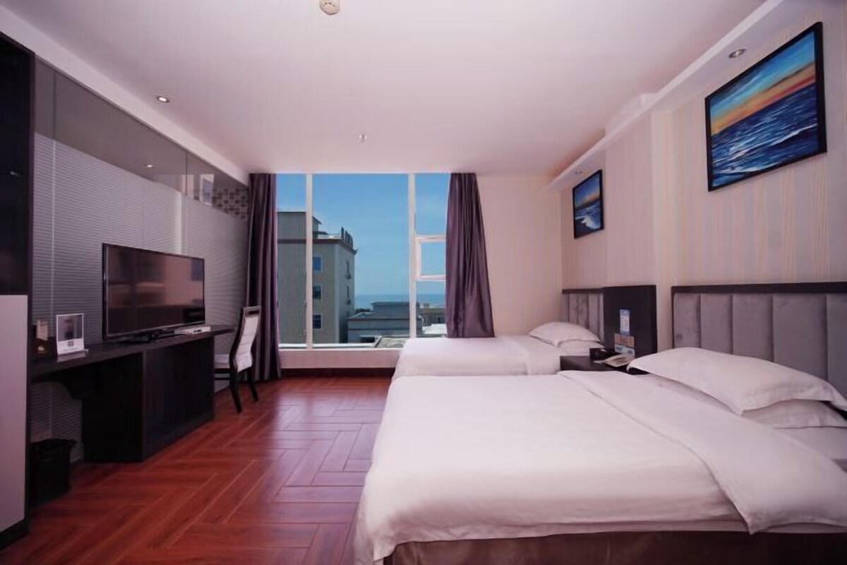 Habitación de hotel con un ventanal grande, un escritorio con televisor, dos camas y un cuadro en la pared.