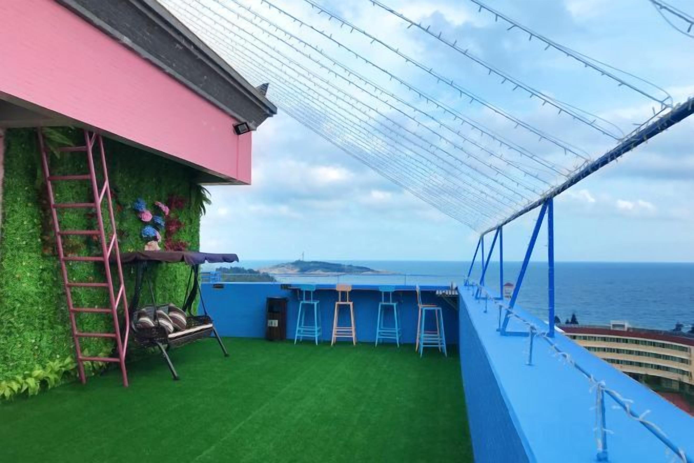 A rooftop terrace with a view of the sea, a green artificial turf, a blue railing, and a pink wall.