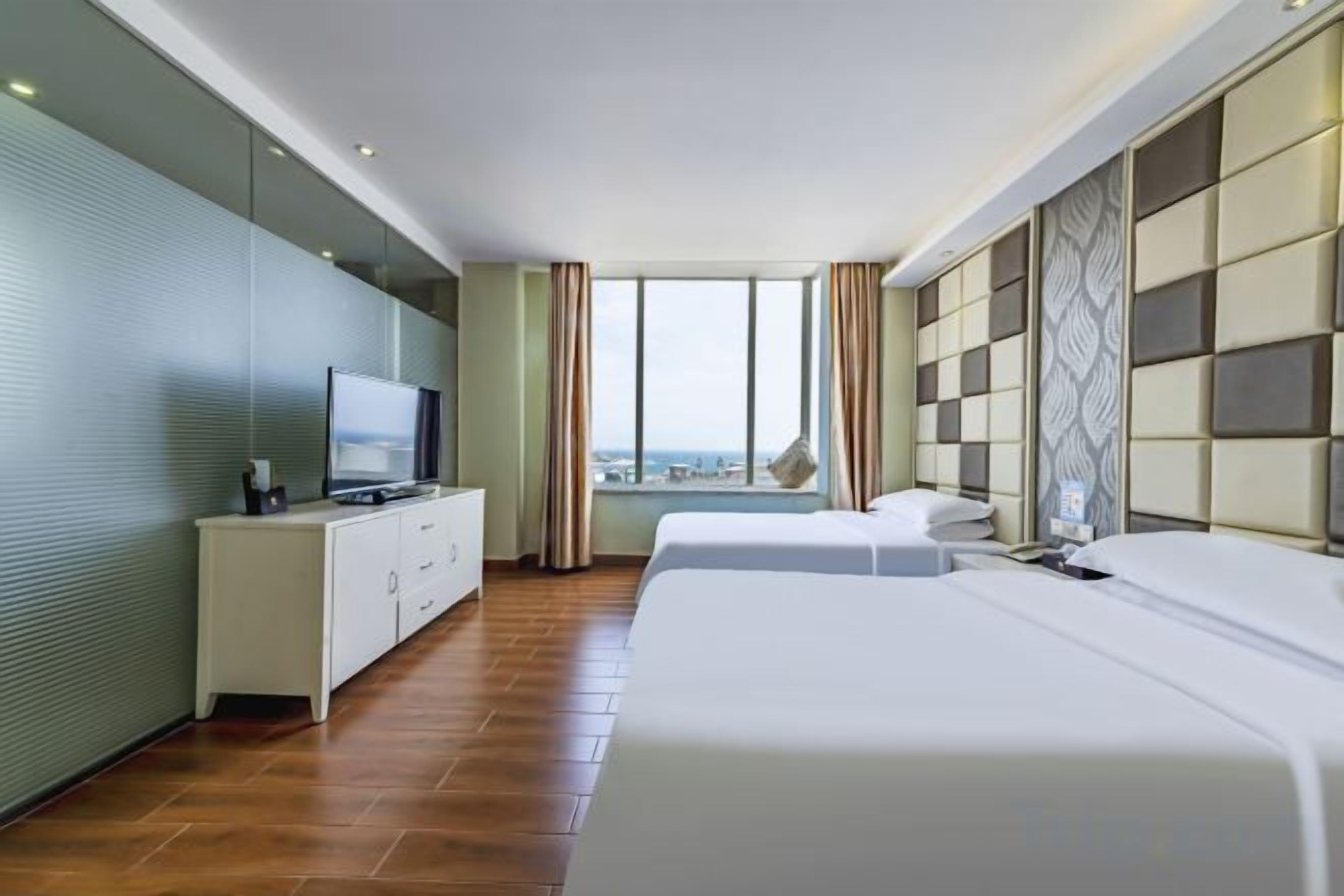 Une chambre d’hôtel avec un lit, un téléviseur posé sur une commode blanche, une grande fenêtre et une vue sur la ville.