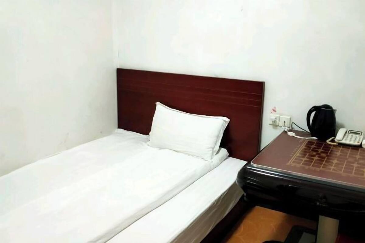 A single bed with a white mattress and a wooden headboard, a small table with a telephone and a kettle, and a wall socket.
