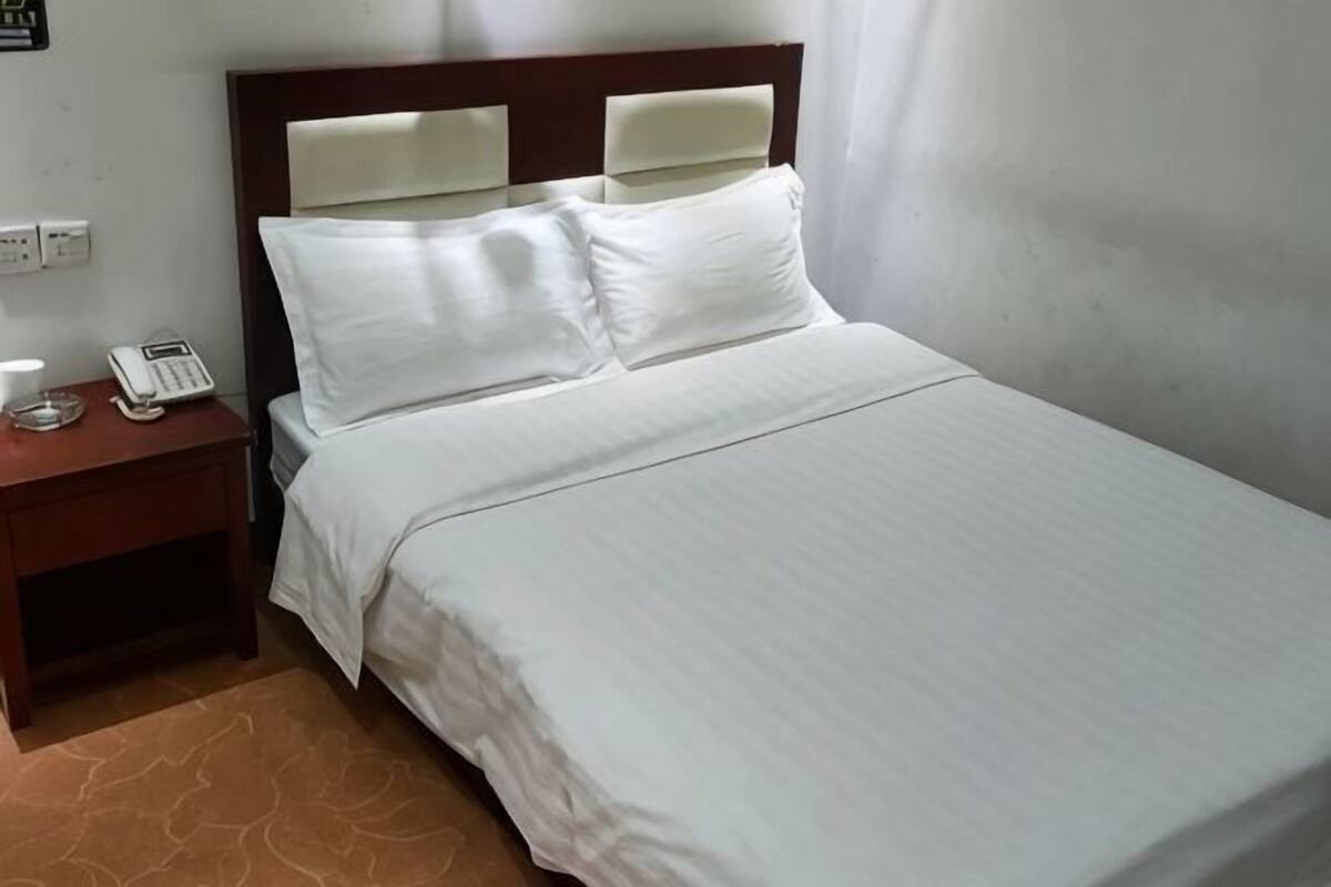 A neatly made bed with white linens, a wooden nightstand with a telephone, and a plain wall with a light switch.