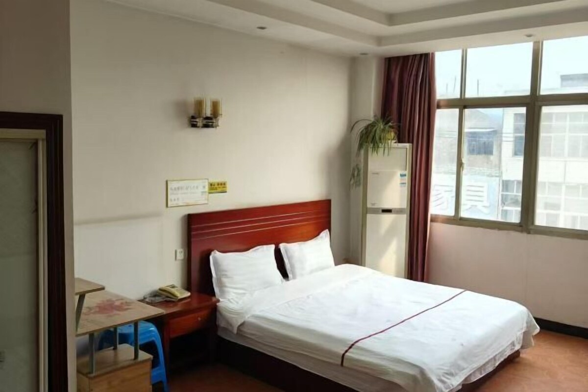 A hotel room with a bed, a desk, a chair, a window, and a plant.