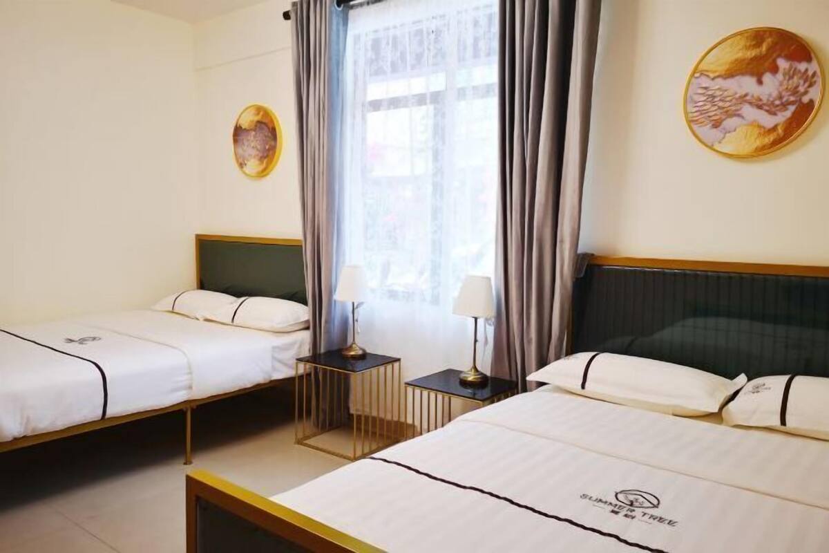 A hotel room with two beds, a window with curtains, a small table with a lamp, and a wall-mounted artwork.