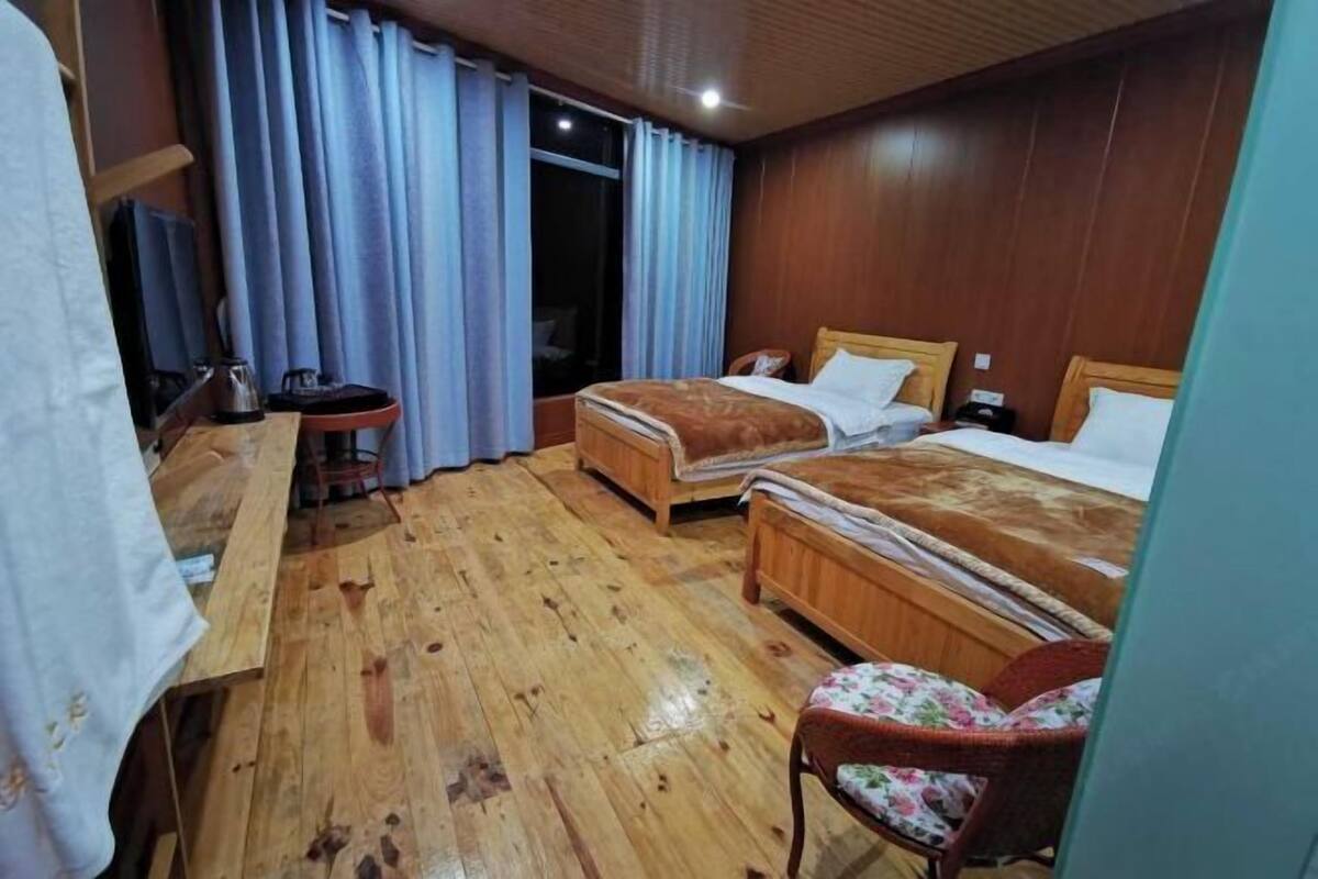 A hotel room with two wooden beds, a small table, a chair, and a window with curtains.