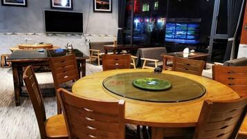 A dining area with a round table, wooden chairs, and a glass-top table. There are multiple screens displaying content in the background.