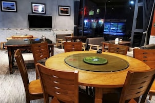 Un comedor con una mesa redonda, sillas de madera y una mesa con cubierta de vidrio. Se observan varias pantallas mostrando contenido en el fondo.