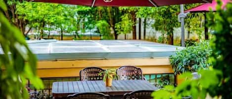 An outdoor seating area with a table and chairs, a large umbrella, and a view of a tennis court.