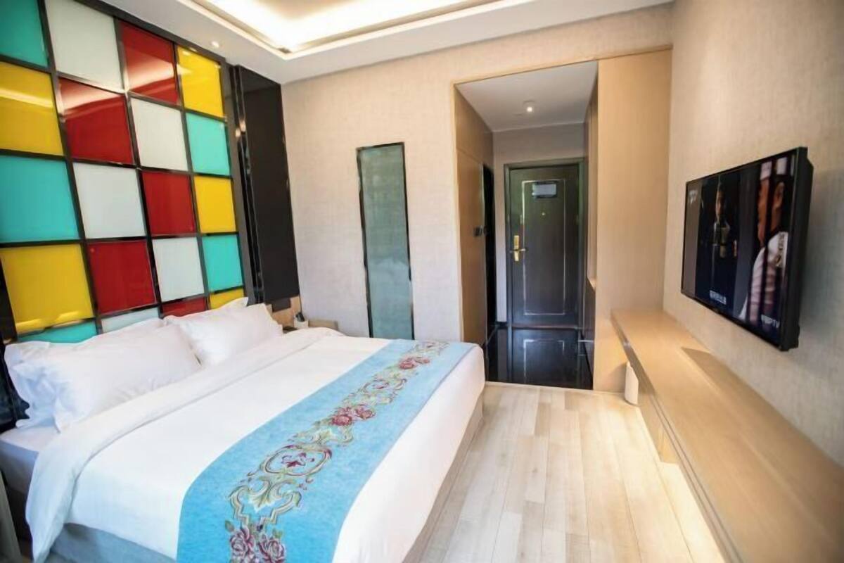 A hotel room with a bed, a TV, a door, and a wall with colorful panels.