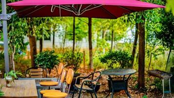 An outdoor seating area with tables and chairs under a large umbrella, surrounded by trees and greenery.