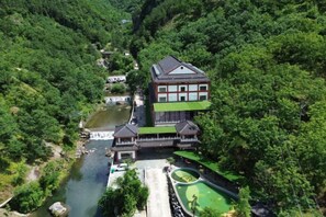 A traditional Japanese building with a multi-tiered structure, surrounded by dense greenery and a river.