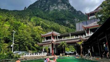 A traditional building with a balcony overlooking a swimming pool area, surrounded by lush greenery and mountains.