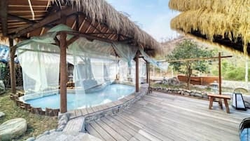 Una terraza de madera con un jacuzzi, una construcción con techo de paja y una zona de piscina.