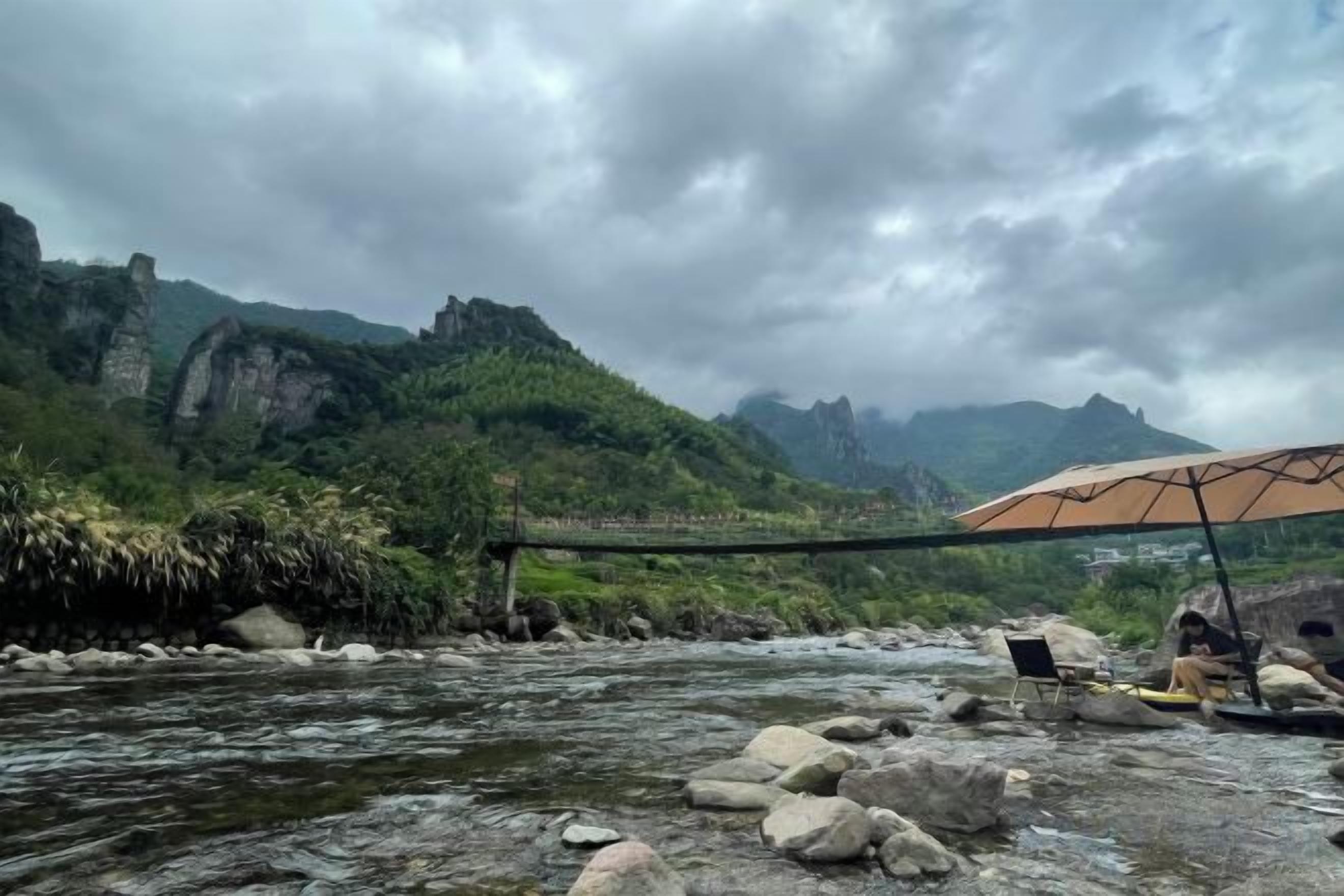 A river with rocks and a bridge, a person under an umbrella, and mountains in the background.