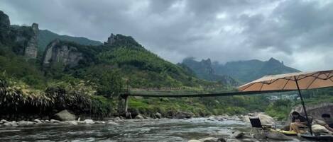 A river with rocks and a bridge, a person under an umbrella, and mountains in the background.