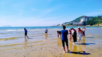 Una playa con gente caminando por la orilla, cielo despejado y un edificio al fondo.