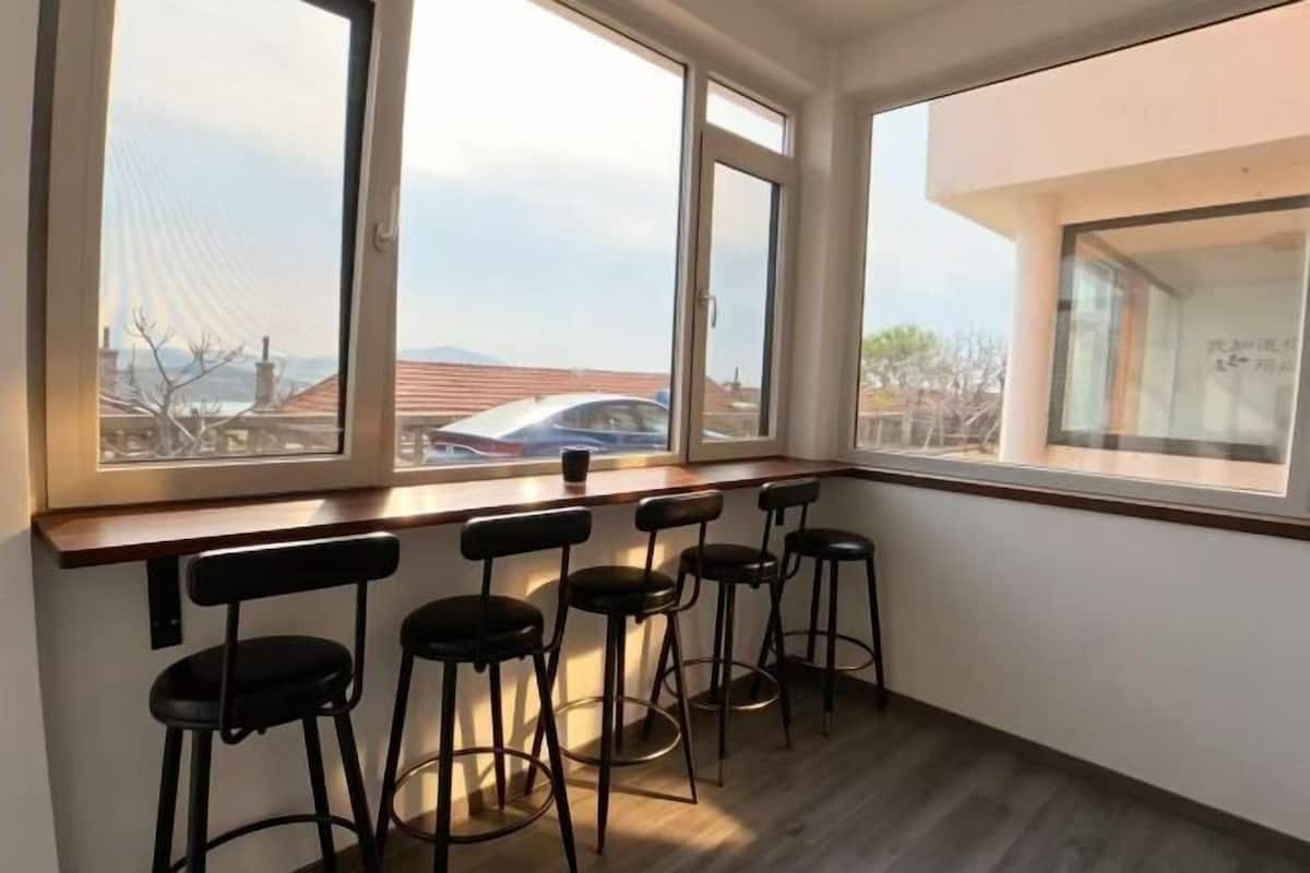 A room with a wooden bar counter and four black bar stools. Large windows provide a view of the outside.