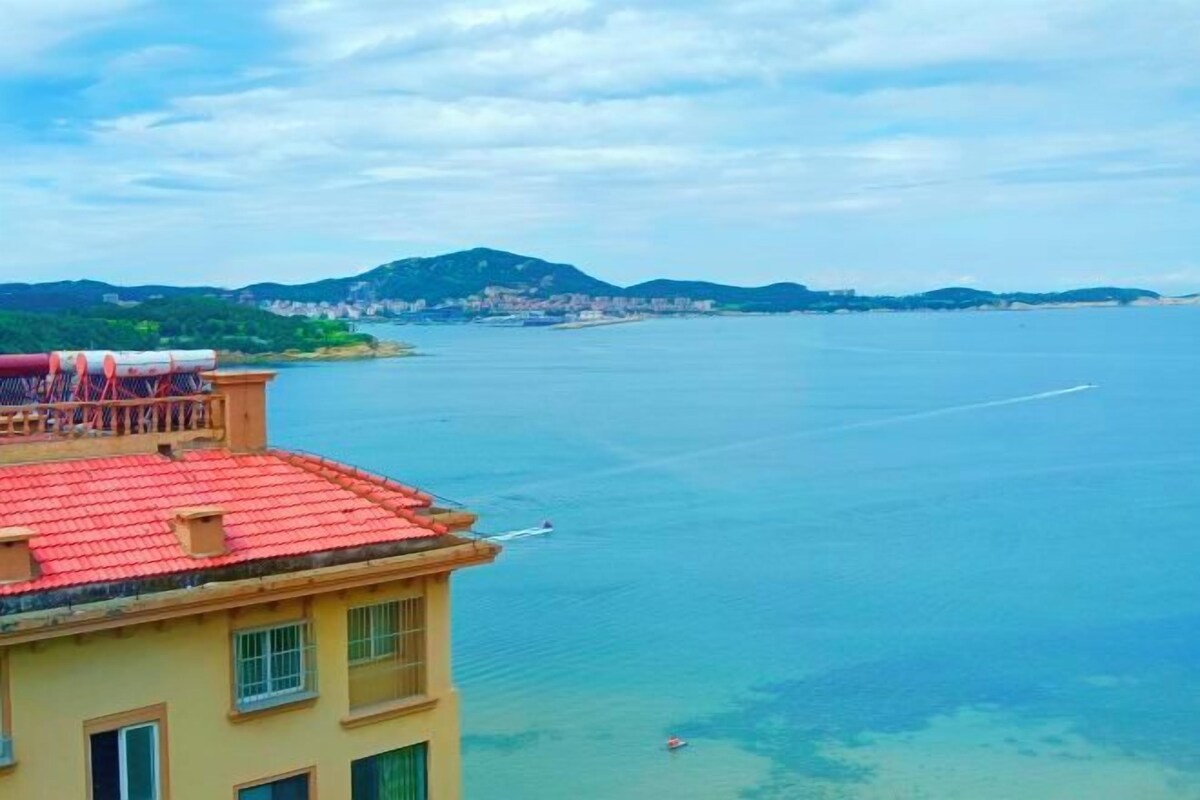 Un edificio costero con techo rojo, con vista al mar tranquilo y un pueblo lejano.