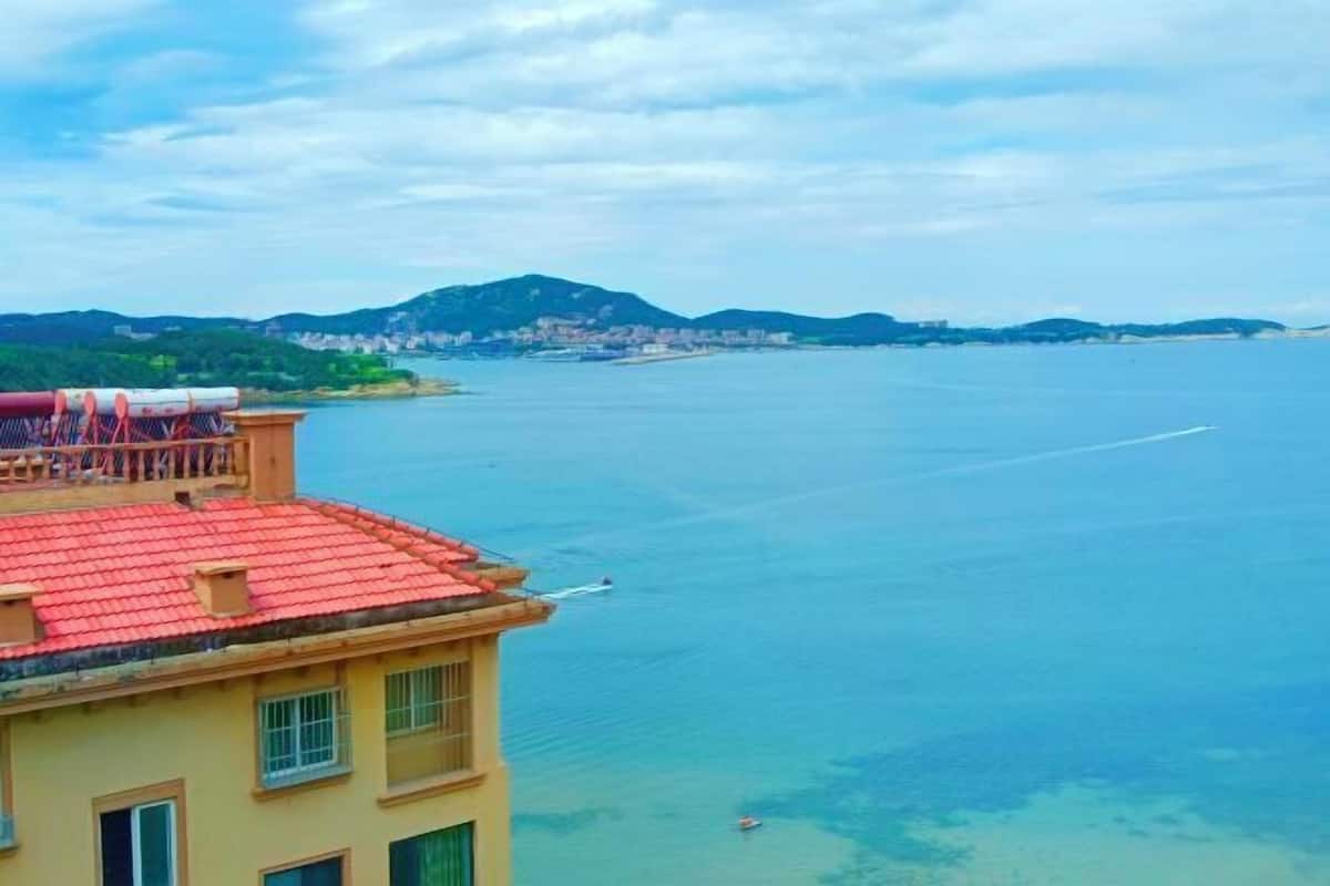 A coastal building with a red-tiled roof overlooking a calm sea and distant hills.