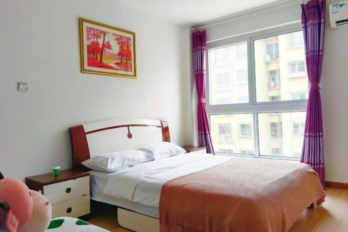 A bedroom with a bed, a nightstand, a window with curtains, and a painting on the wall.