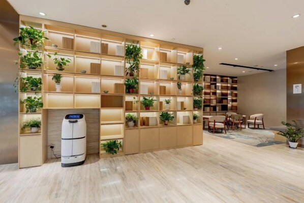 A hotel lobby with a large shelving unit displaying potted plants, a seating area with chairs, and a white cylindrical object on the floor.