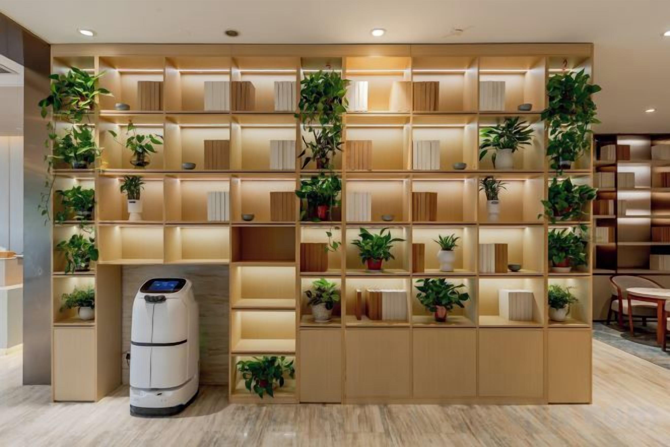 A wall with multiple shelves containing books and potted plants, a white device on the floor, and a wooden structure on the left.
