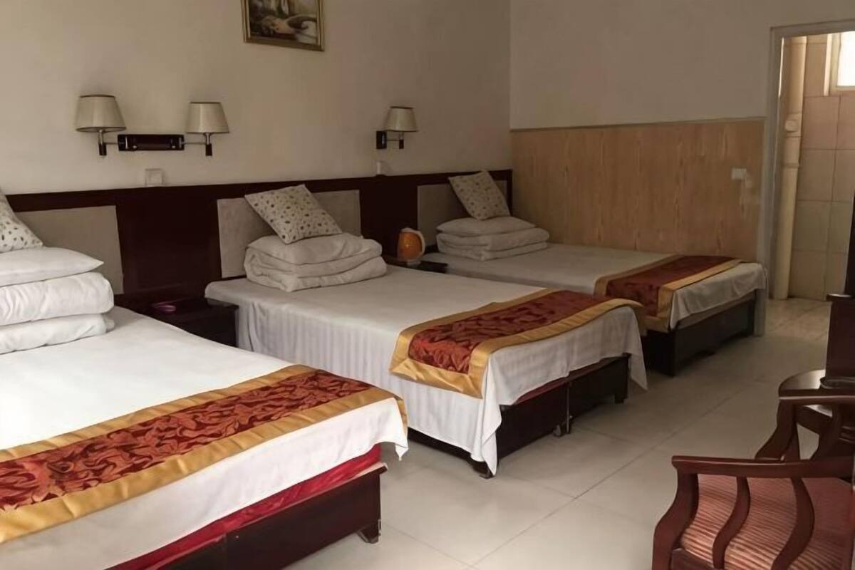 A hotel room with three beds, each with a white bedspread and a red and gold decorative border. There are two wall-mounted lamps on the wall above the beds.