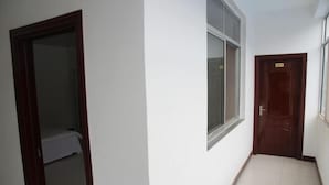 Un couloir étroit, avec une porte au fond, une fenêtre sur la droite et une pièce partiellement visible, où l’on aperçoit un lit.