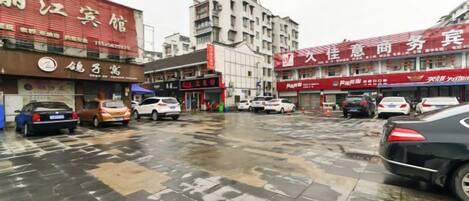 A parking lot with multiple cars, a wet ground indicating recent rain, and buildings with commercial signage in a city setting.