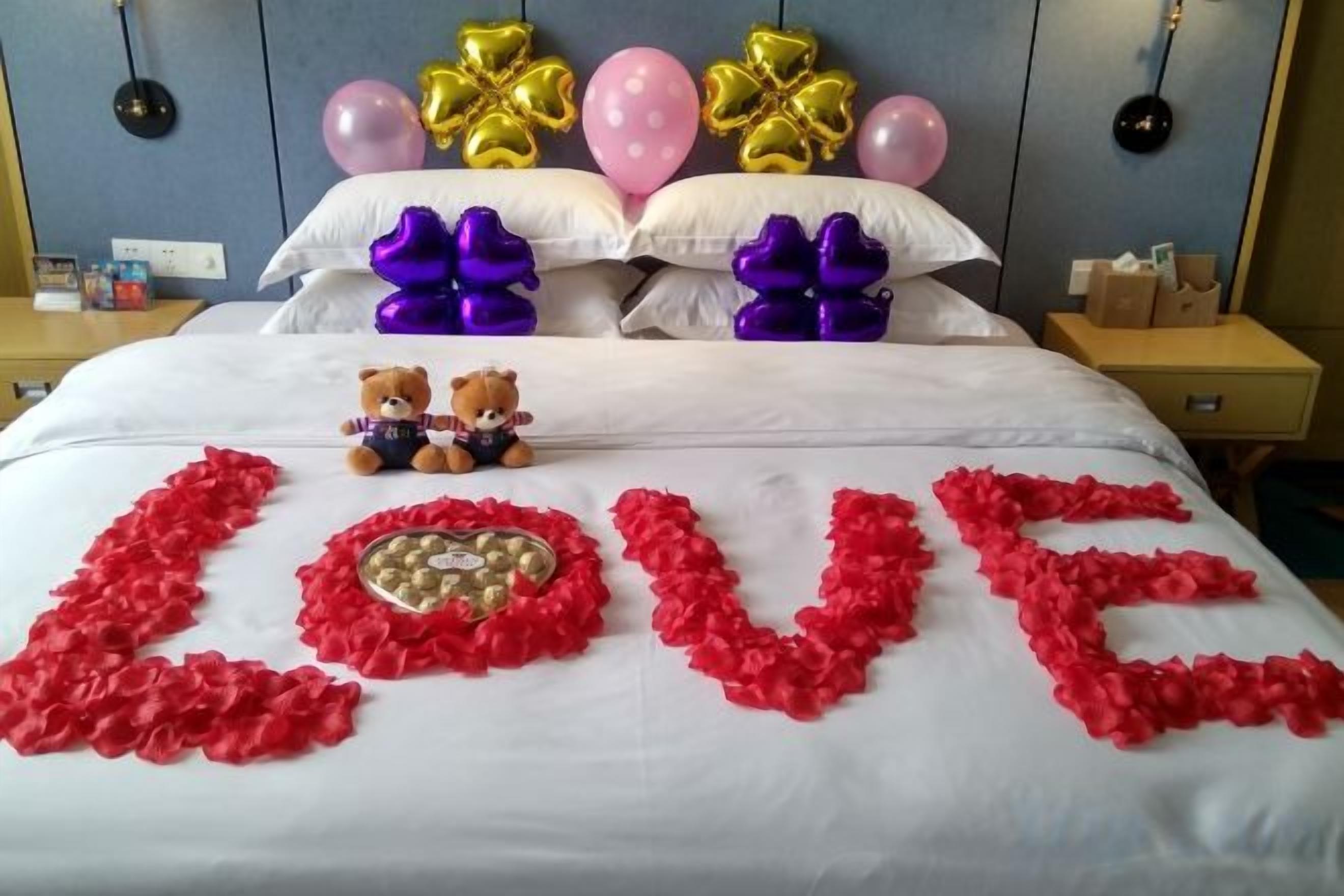 A bed with a 'LOVE' decoration made of red rose petals, two teddy bears, and balloons on the headboard.