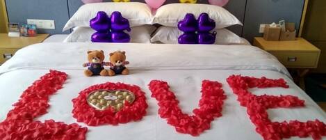 A bed with a 'LOVE' decoration made of red rose petals, two teddy bears, and balloons on the headboard.