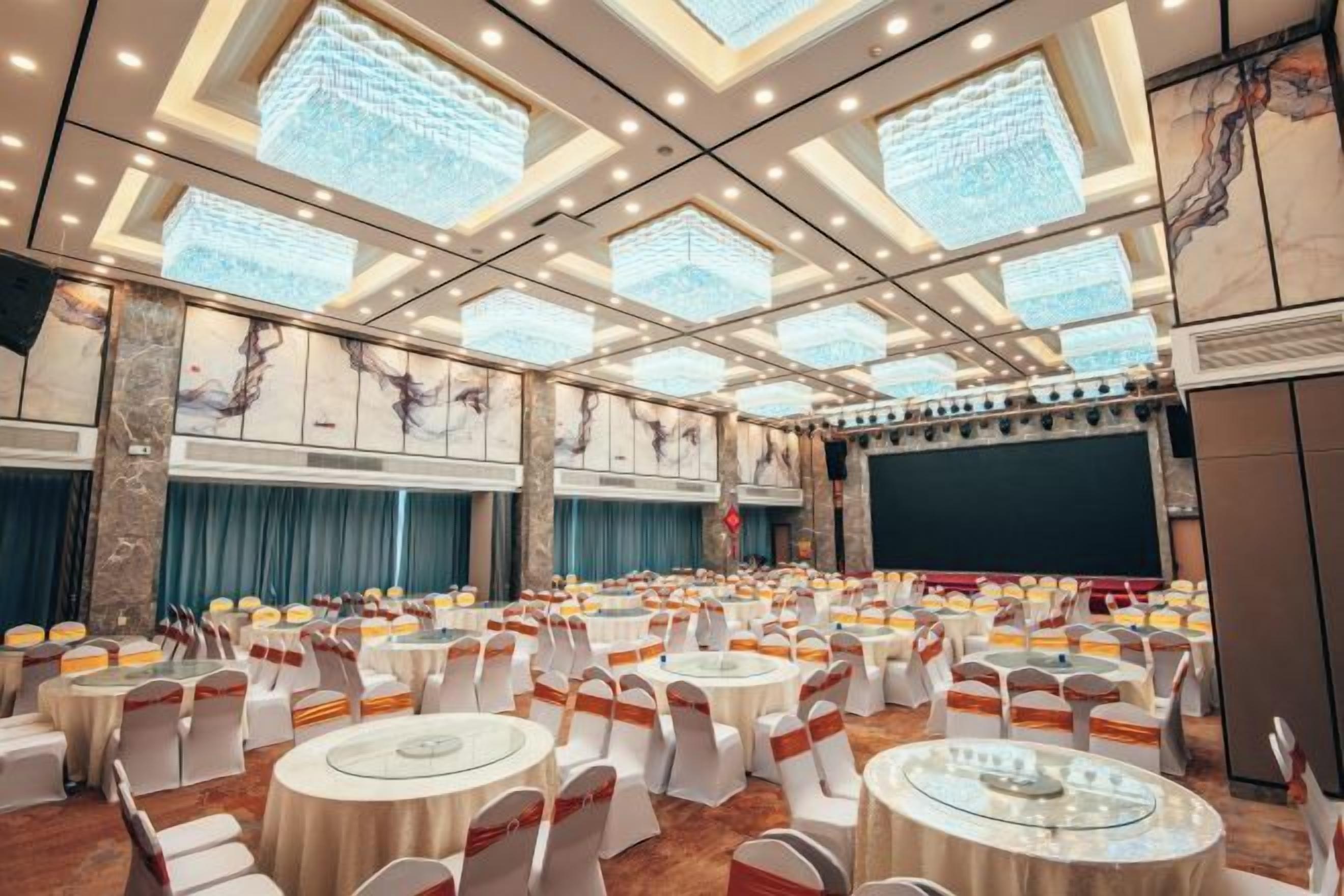 Un salón de banquetes con mesas redondas, sillas blancas y un escenario.