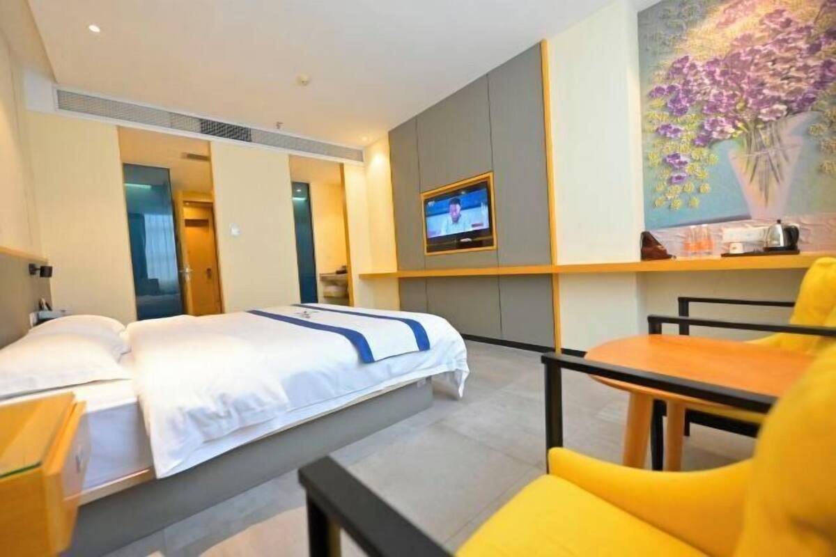 A hotel room with a bed, a TV mounted on the wall, a painting of flowers, and a small table with a chair.