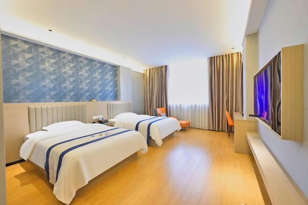 A hotel room with two beds, a large window with curtains, a flat-screen TV mounted on the wall, and a wooden floor.