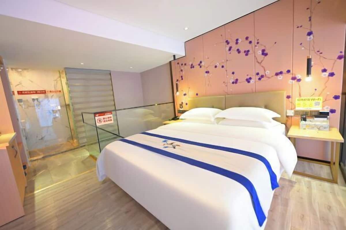 A modern hotel room with a large bed, a glass partition, and a decorative wall with purple flowers.