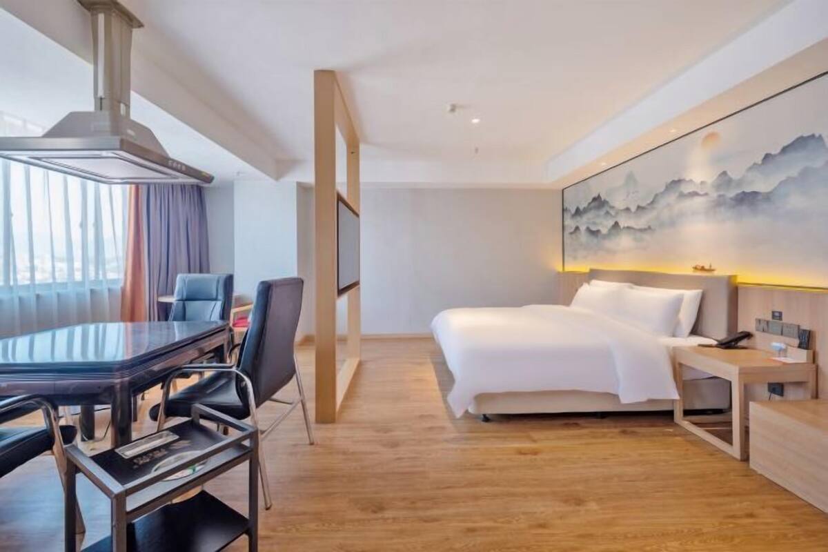 A hotel room with a wooden floor, a large bed, a desk with chairs, a painting on the wall, and a view of the city through the window.