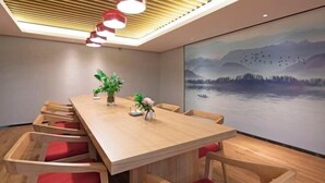 Une salle de conférence moderne, dotée d’une table en bois et de chaises rembourrées de couleur rouge, d’un grand tableau représentant un paysage montagneux, et d’un plafond orné de lambris en bois et de luminaires suspendus.
