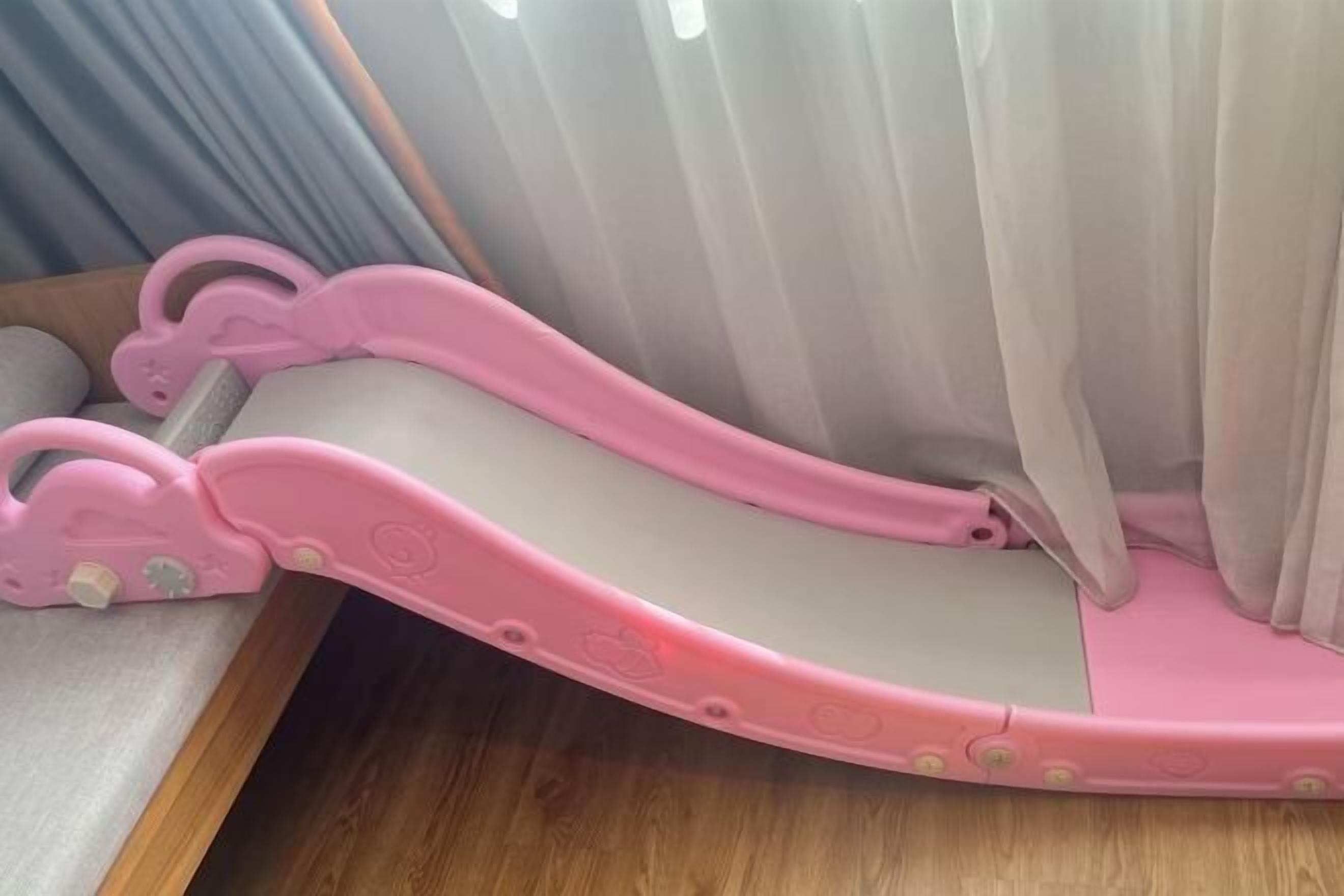 A pink baby walker with a curved design, positioned near a window with white curtains.