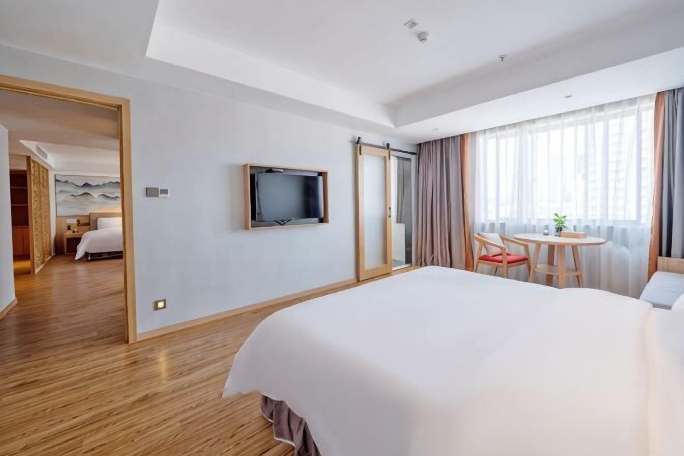 Habitación de hotel con una cama grande, un televisor montado en la pared, zona de comedor con mesa y sillas, y vista a la ciudad a través de amplios ventanales.