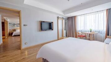 Habitación de hotel con una cama grande, un televisor montado en la pared, zona de comedor con mesa y sillas, y vista a la ciudad a través de amplios ventanales.