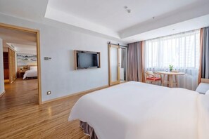 Habitación de hotel con una cama grande, un televisor montado en la pared, zona de comedor con mesa y sillas, y vista a la ciudad a través de amplios ventanales.