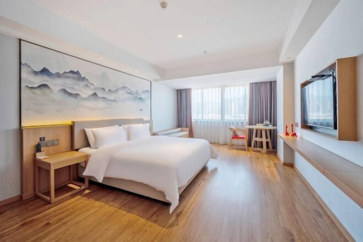 A modern hotel room with a large bed, a bedside table, a desk, and a large window.