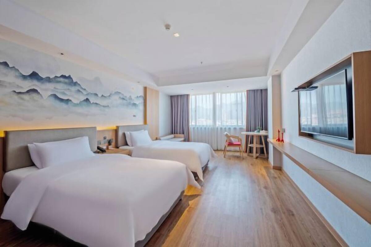 A hotel room with two beds, a desk, a chair, a large window, and a painting on the wall.
